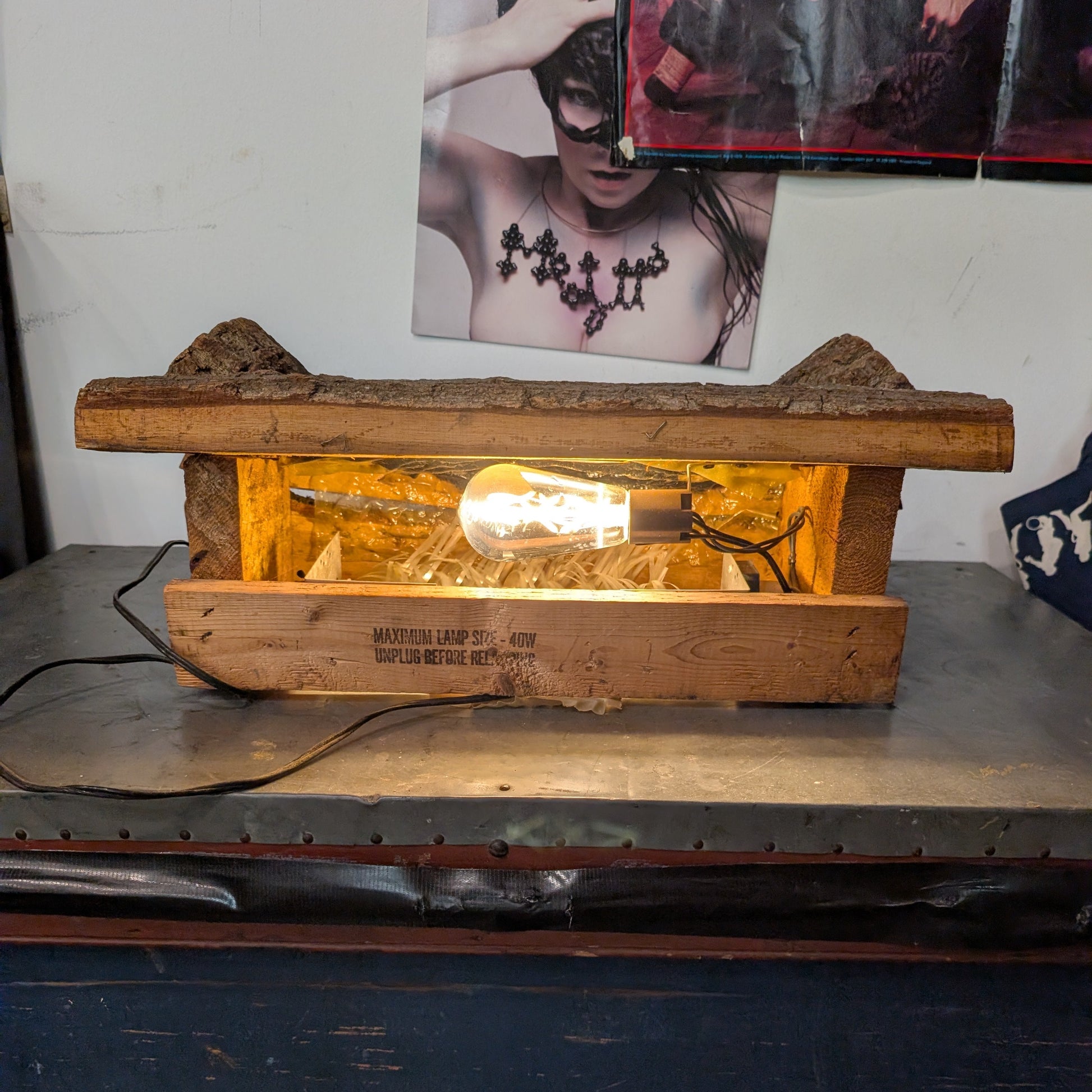 A rustic wooden box lamp with a glowing light bulb inside, placed on a dark metal surface with a wall and posters in the background.